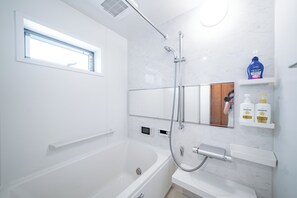 A modern bathroom with a bathtub, a shower, a vanity with a mirror, and a window above the bathtub. - Tokyo Home Base Haneda (Tokyo)