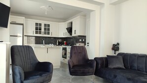 Ein modernes Wohnzimmer mit einem dunkelblauen Sofa, einem passenden Sessel und einem kleineren Stuhl. Im Hintergrund ist eine Küche mit weißen Schränken und einem schwarzen Herd zu sehen.