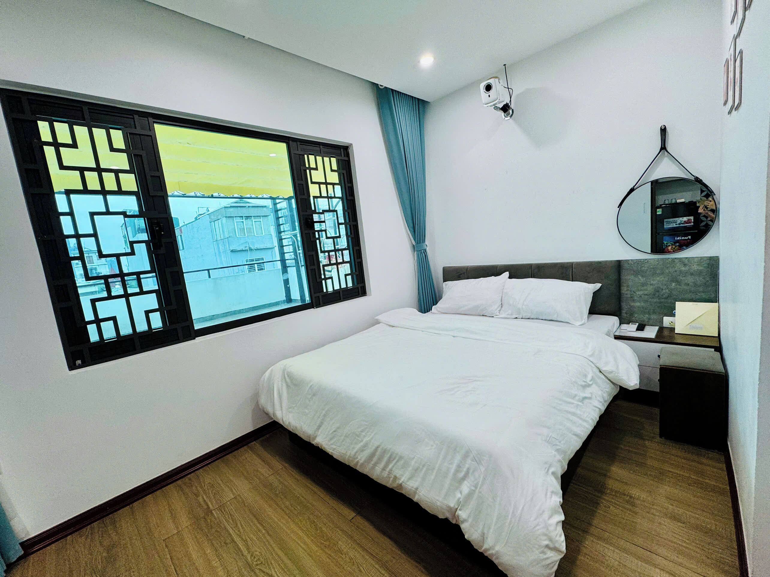 Habitación de hotel con cama, mesita de noche, espejo y ventana con vista a edificios.