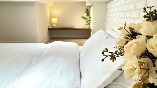 A hotel room with a white brick wall, a bed with white linens, a bedside table with a lamp, and a bouquet of white flowers. - Ly Home Tay Ho Ha Noi  (Hanoi)