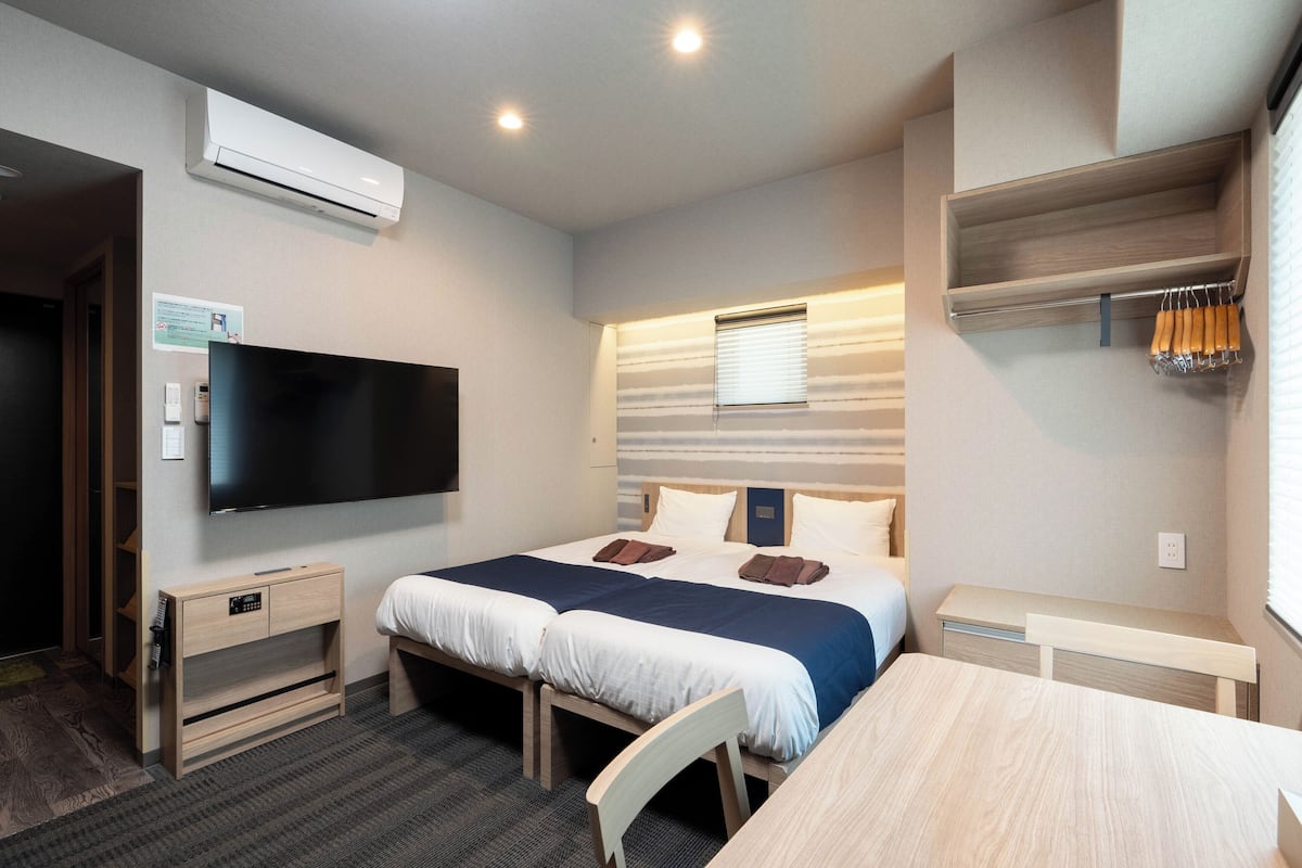 A hotel room with two beds, a TV, a desk, and a window with blinds.