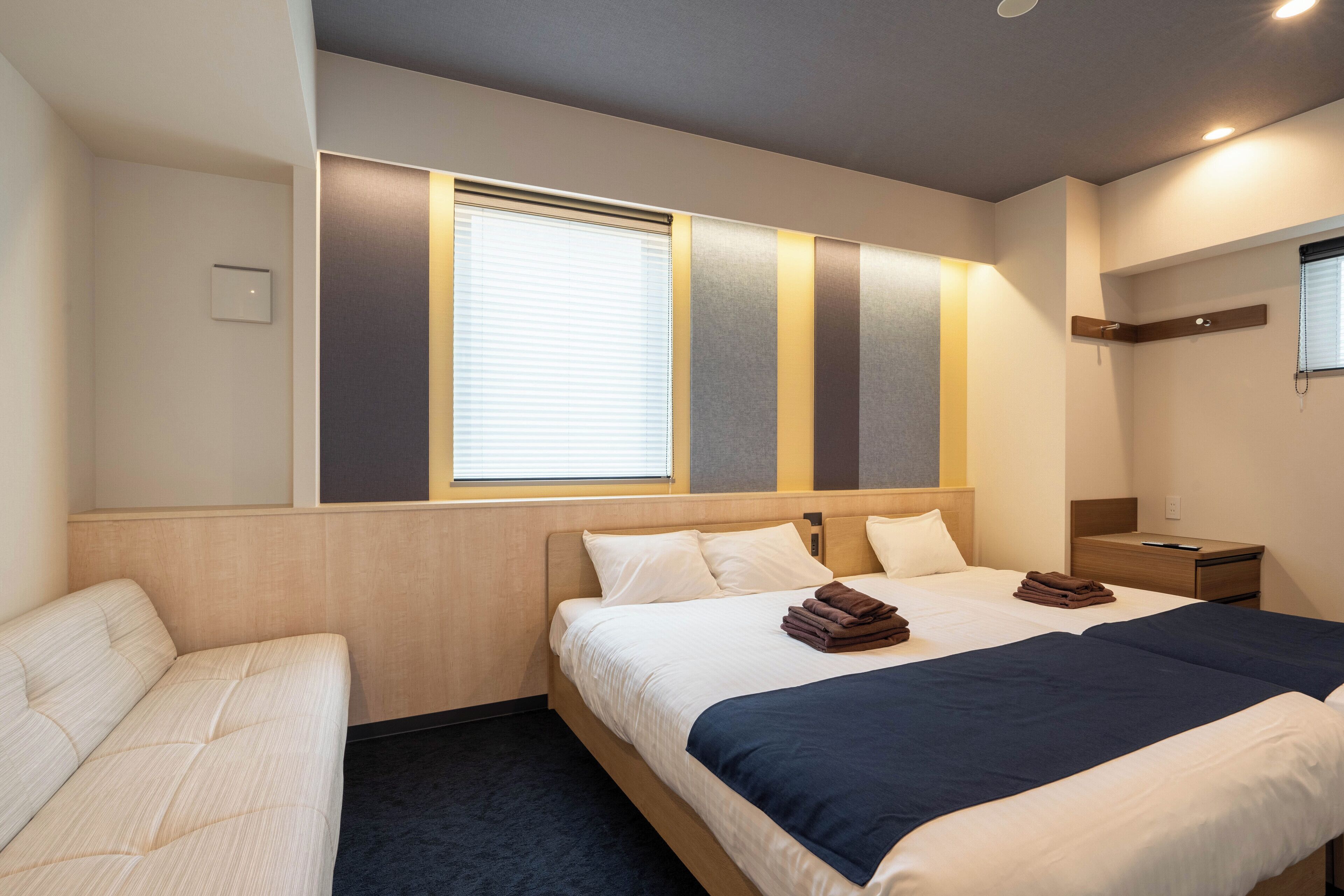 A hotel room with a large bed, a bedside table, a nightstand, a chair, and a window with blinds.