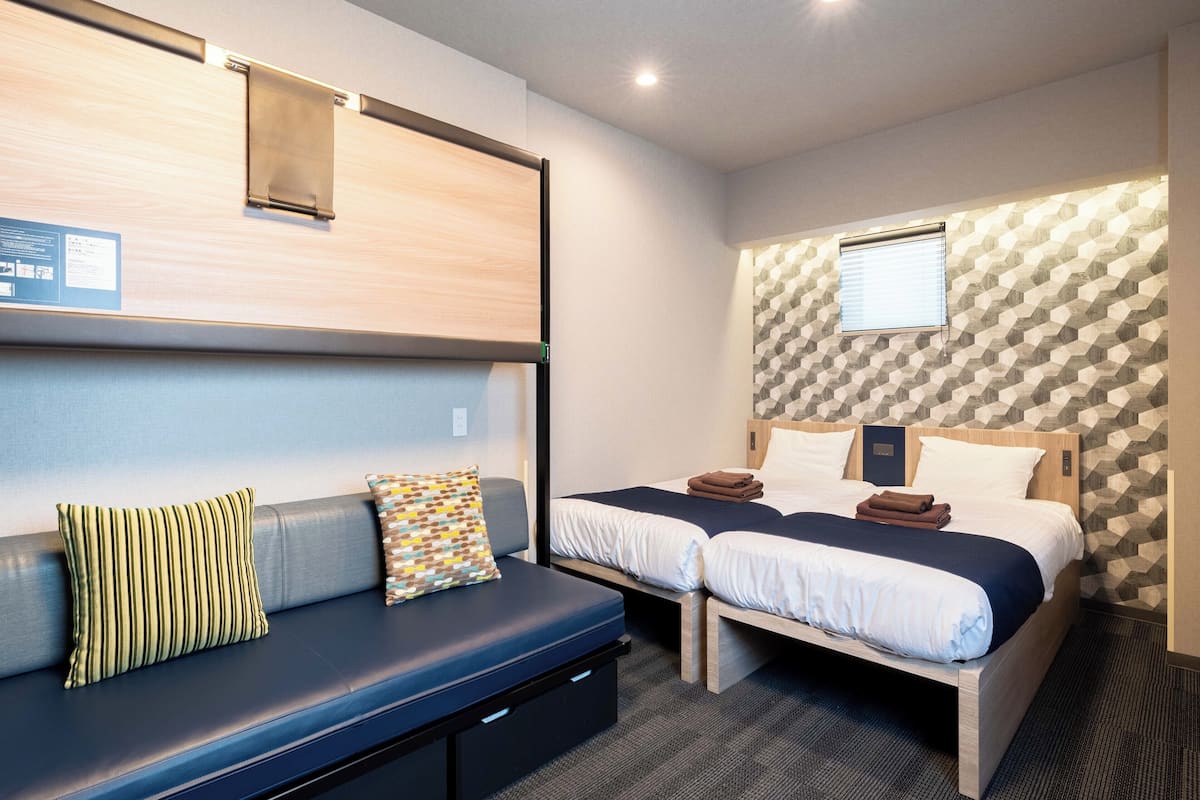 A hotel room with a sofa, two beds, a window, and a patterned wall.