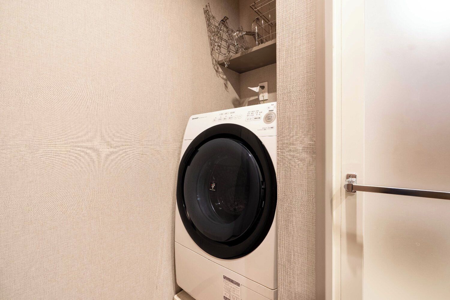 Eine Waschmaschine mit Frontlader in einem Waschraum mit hellbeige Wänden und einem Metallregal darüber.