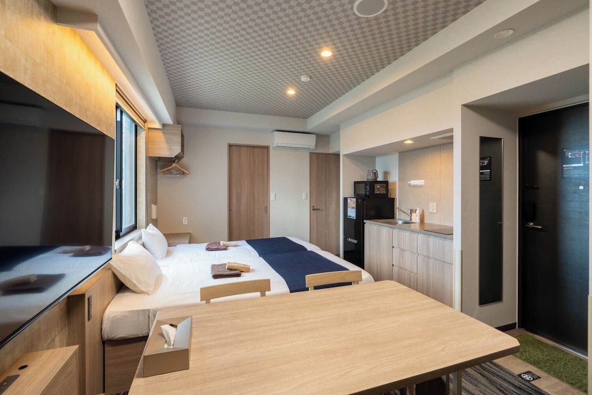 A compact hotel room with a bed, bedside table, a small kitchenette, and a view through a window.