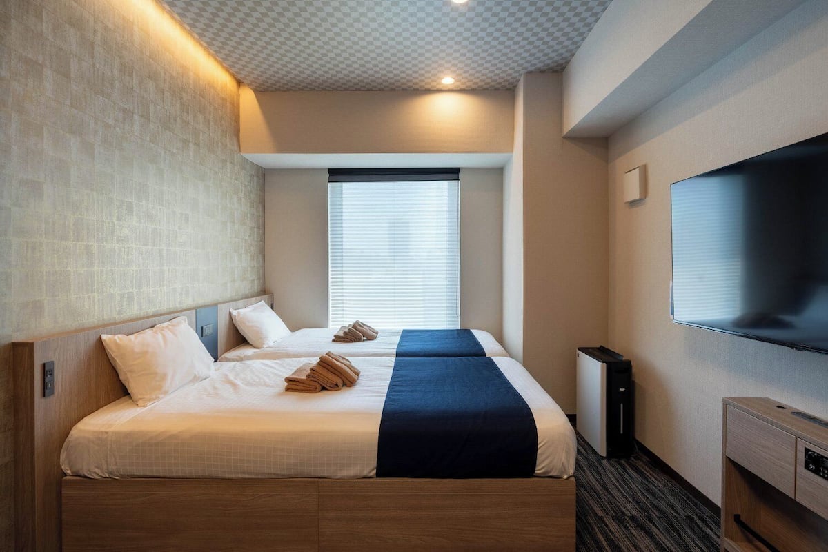 A hotel room with two beds, a flat-screen TV, and a window with blinds.