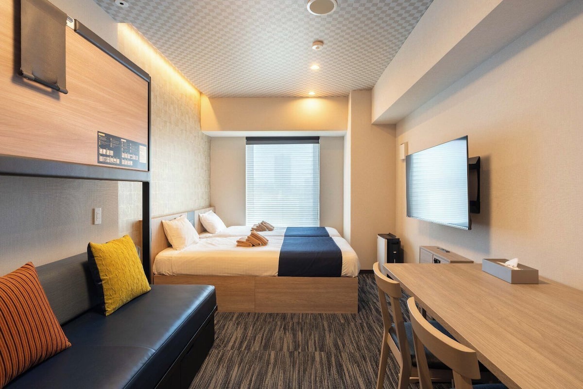 A modern hotel room with a bed, a desk, a TV, and a seating area.