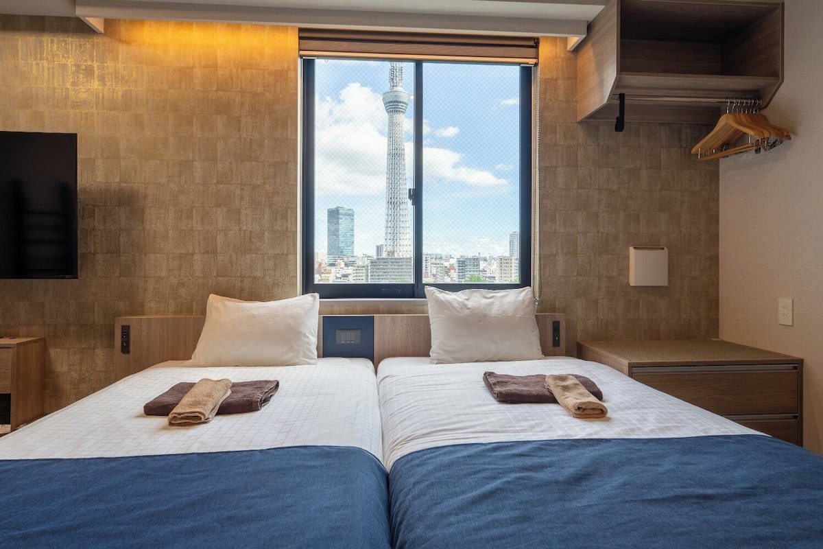 A hotel room with two beds, a large window with a city view, a TV, and a wooden wardrobe.