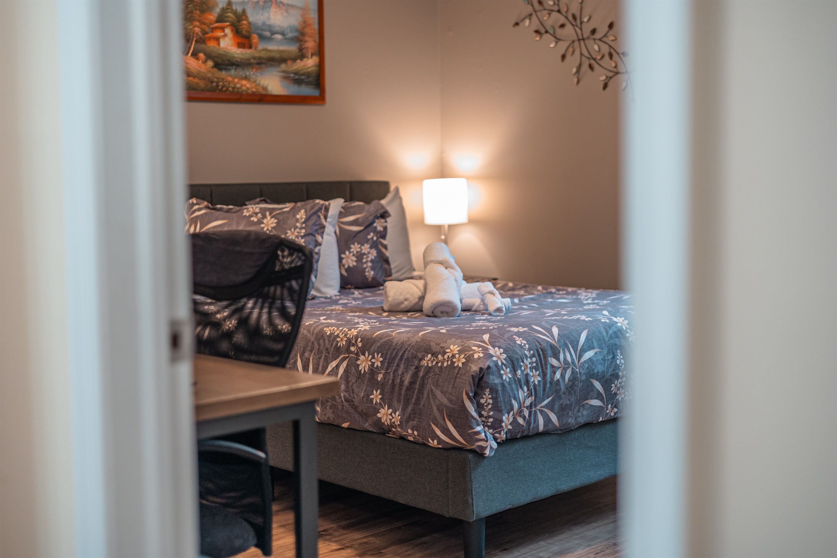 En pænt redt seng med et blomstret sengetæppe, en senget lampe og en blød legetøj på sengen.