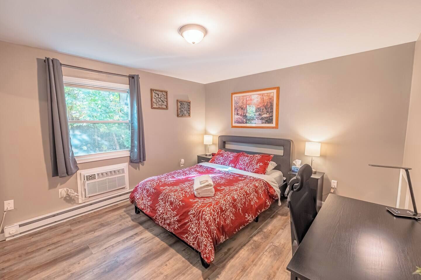 Quarto com cama, duas mesas de cabeceira, escrivaninha, cadeira, janela com cortinas e radiador.