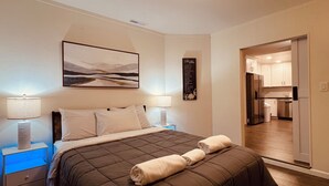 Een slaapkamer met een bed, twee nachtkastjes, een wanddecoratie en een keuken die door een open deur zichtbaar is.