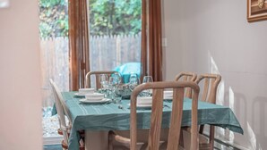 Una mesa de comedor dispuesta con platos, vasos y cubiertos, cubierta con un mantel verde azulado, en una habitación con sillas de madera y vistas a follaje exterior.