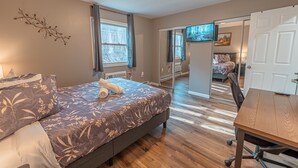 Une chambre à coucher avec un lit, un bureau et une chaise. Il y a une fenêtre avec des rideaux, un élément décoratif fixé au mur et une porte qui donne sur une autre pièce.