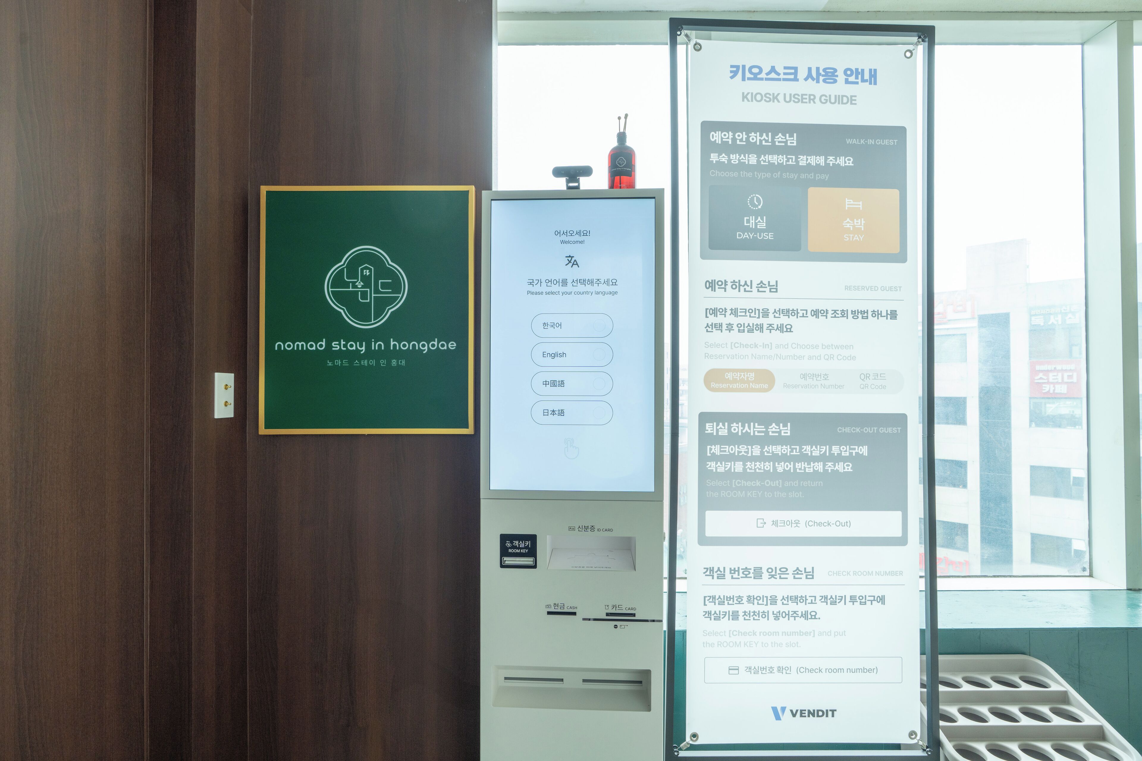 Ein Hotel-Rezeptionsbereich mit einer digitalen Kiosk-Station für den Gäste-Check-in, einem gerahmten Schild mit der Aufschrift 'nomad stay in Hongdae' und einem Fenster mit Blick auf Gebäude.