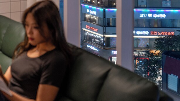 Une femme est assise sur un canapé vert dans une pièce d’où l’on aperçoit un bâtiment dont les enseignes lumineuses sont rédigées dans une langue étrangère.
