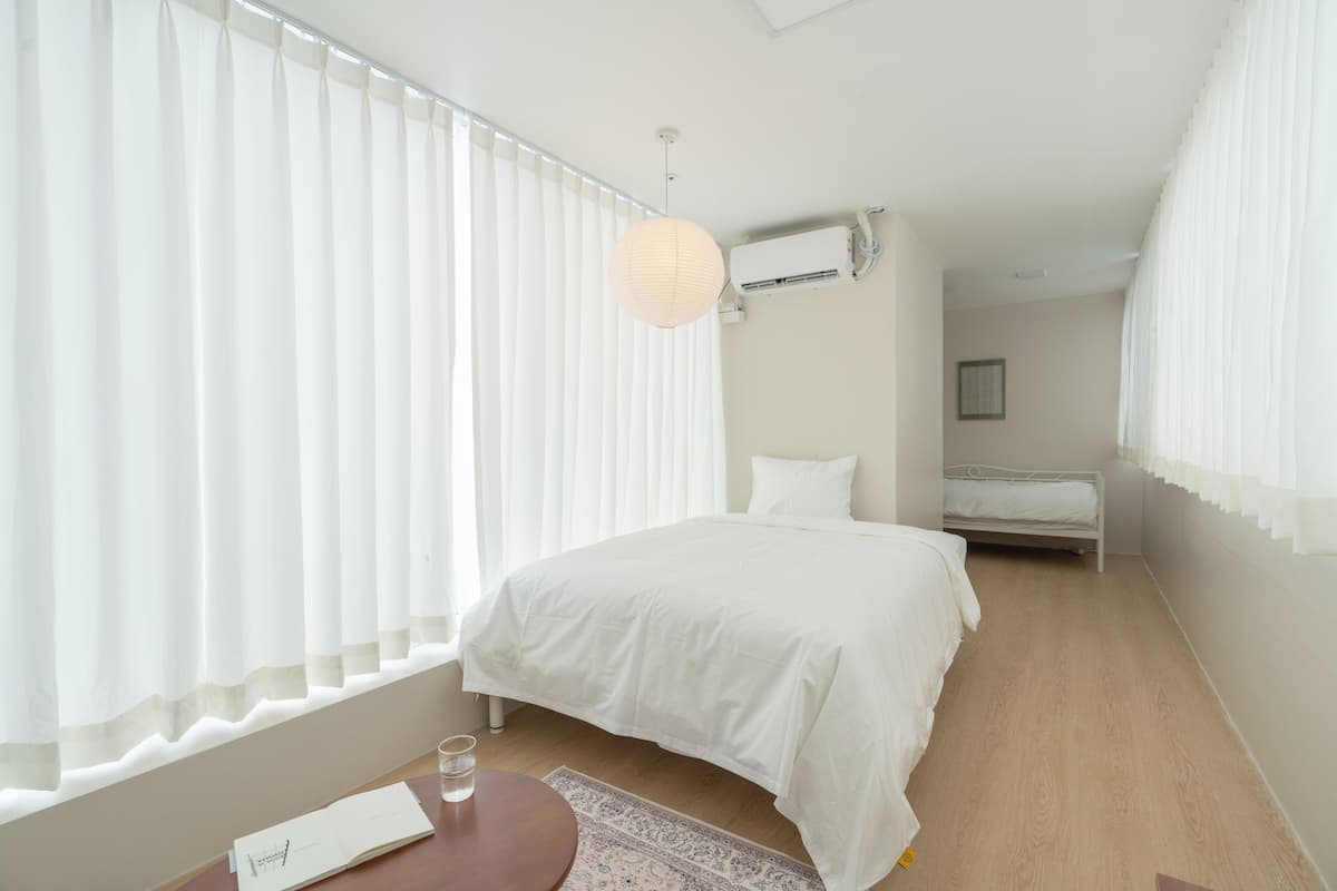 A bedroom with a bed, a bedside table with a book and glass, a hanging light, and a window with blinds.