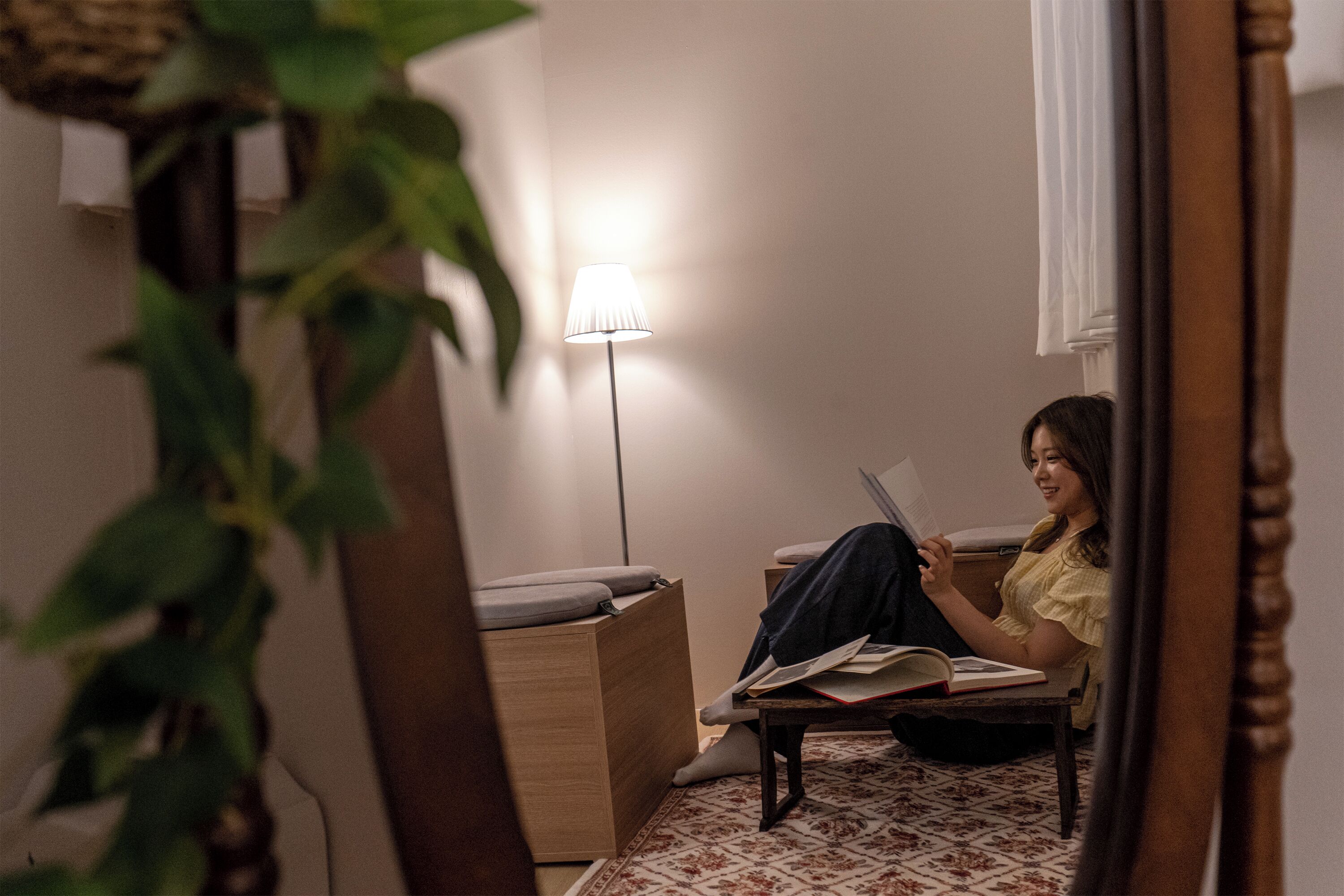A person sitting on a chair reading a book in a room with a lamp, a small table, and a plant.