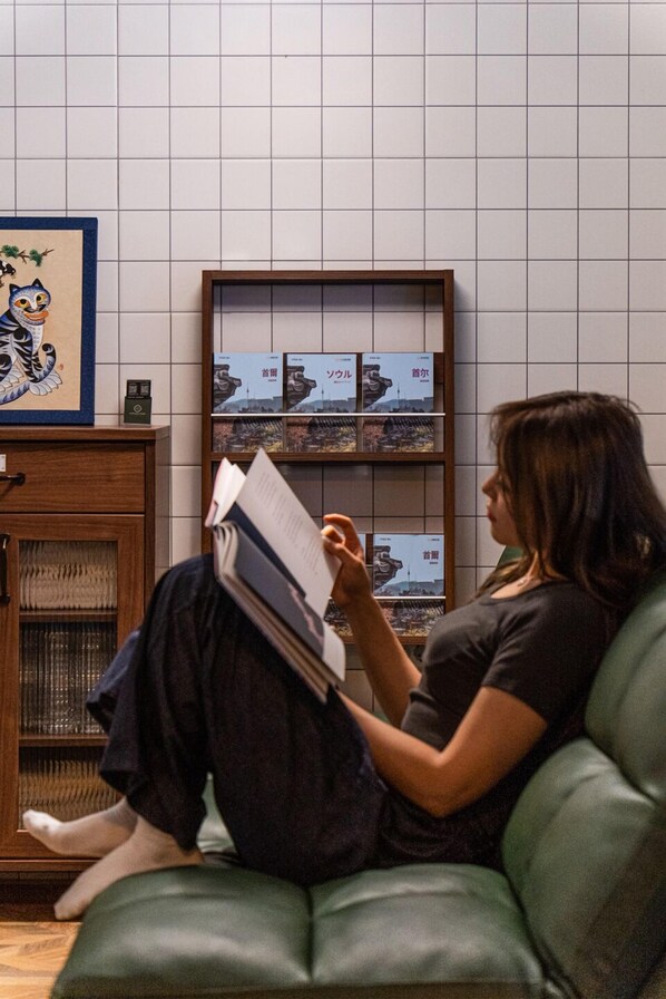 A person sitting on a green couch reading a book in a room with a tiled wall and a bookshelf with books and a framed picture.