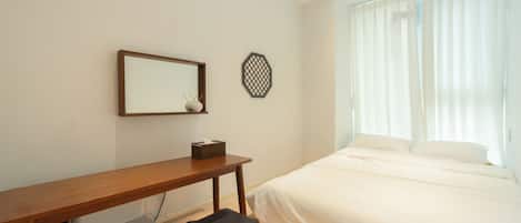 A hotel room with a wooden desk, a small stool, a bed with white linens, a wall-mounted mirror, and a decorative wall piece.