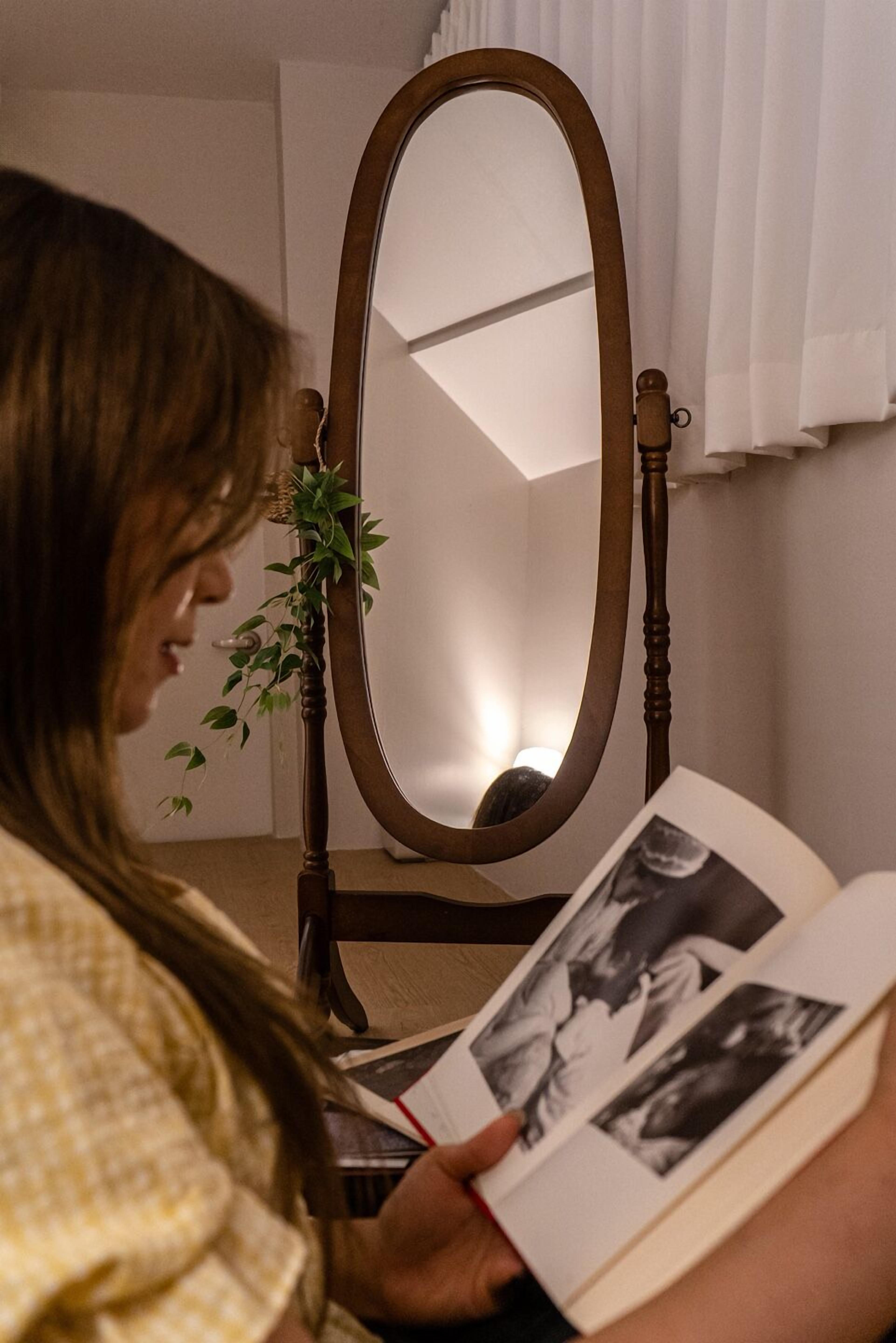 A person reading a book in a room with a wooden chair and a mirror.