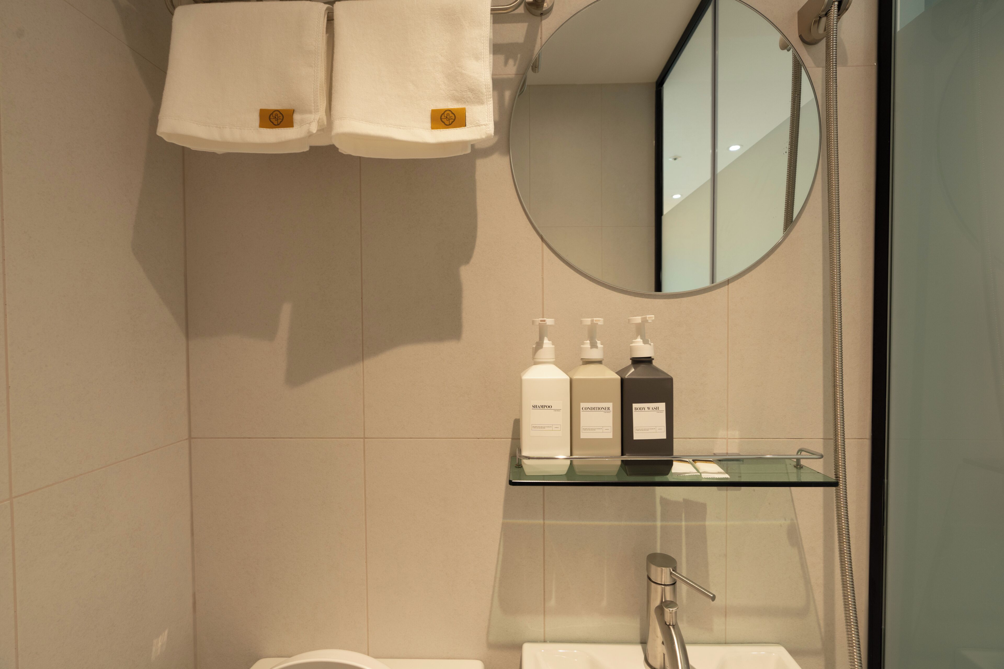 A bathroom with a glass shelf holding toiletries, a round mirror, and two towels hanging on a towel rack.