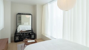 Une chambre d’hôtel avec un grand lit, une commode avec un miroir, une petite table et une grande fenêtre avec des rideaux légers.