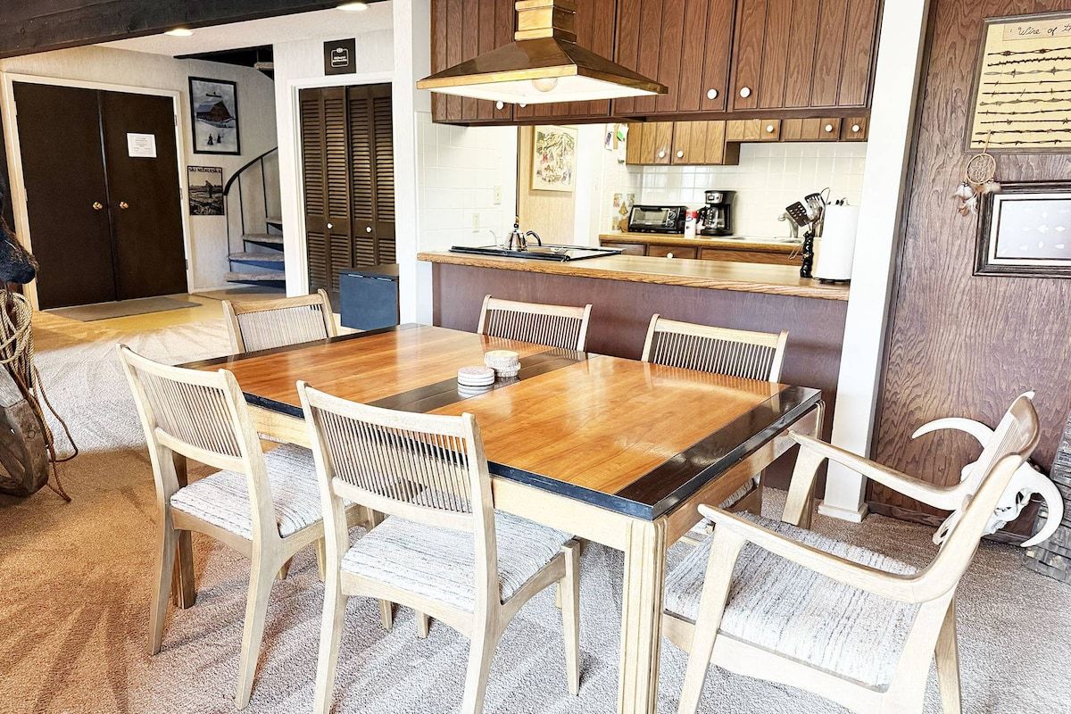 A dining area with a wooden table and chairs, a kitchen in the background, and a staircase leading to another level.