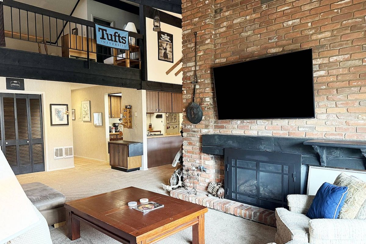 A loft-style living area with a fireplace, a television mounted on the wall, and a sign reading 'Tufts University'.