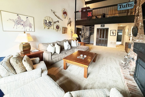 A spacious living room with a sofa, coffee table, and fireplace. A staircase leads to an upper level with a sign reading "Tufts University".