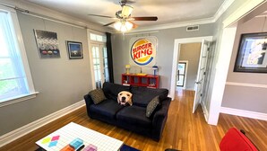 Een woonkamer met een donkergrijze bank, een tafel met kleurrijke geometrische vormen, een plafondventilator en een Burger King bord aan de muur.