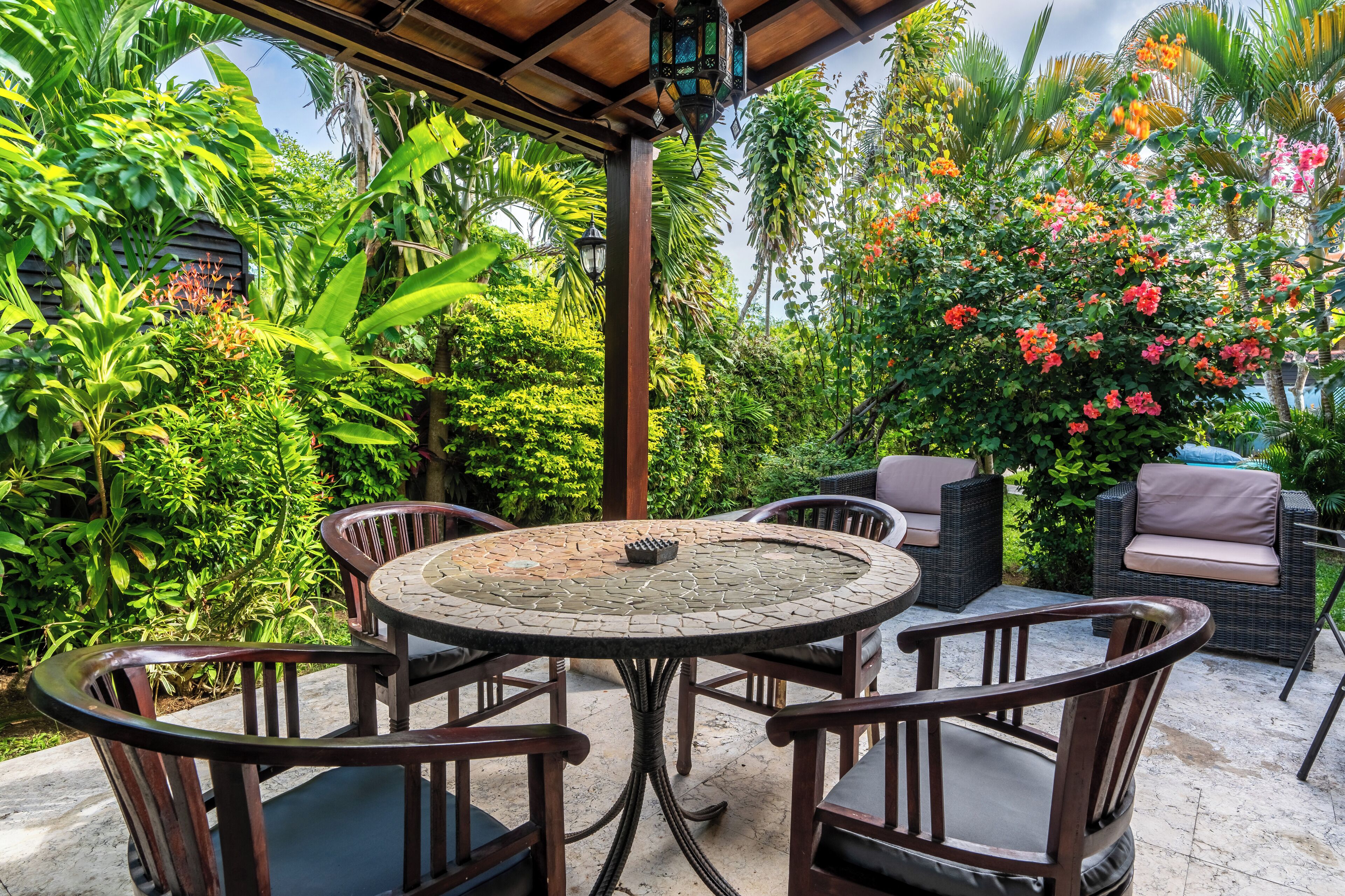 A covered patio with a round table and chairs, surrounded by lush greenery and flowering plants.