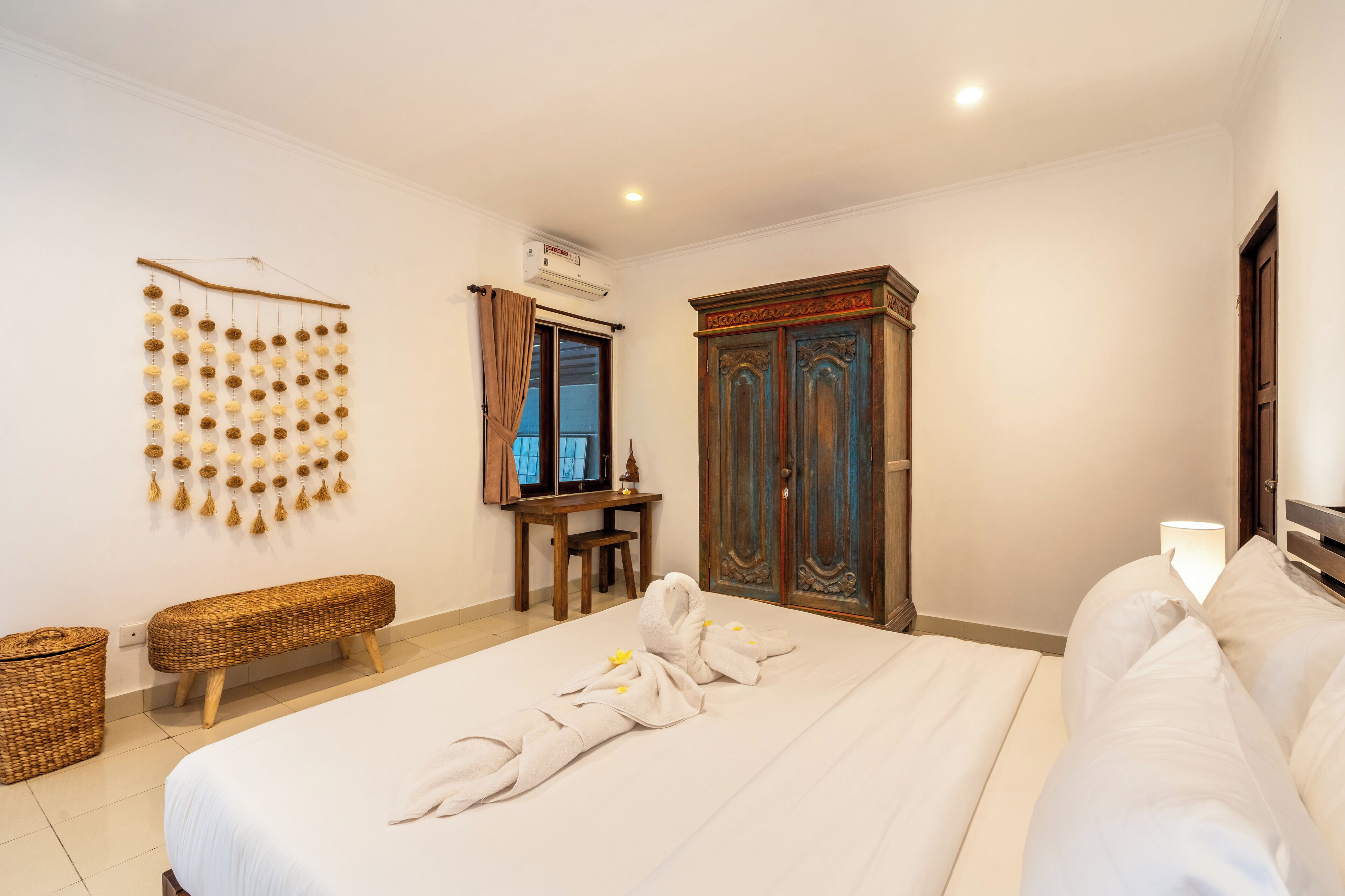 A hotel room with a large bed, a wooden wardrobe, a small desk with a chair, a wall-mounted air conditioner, and a decorative wall hanging.
