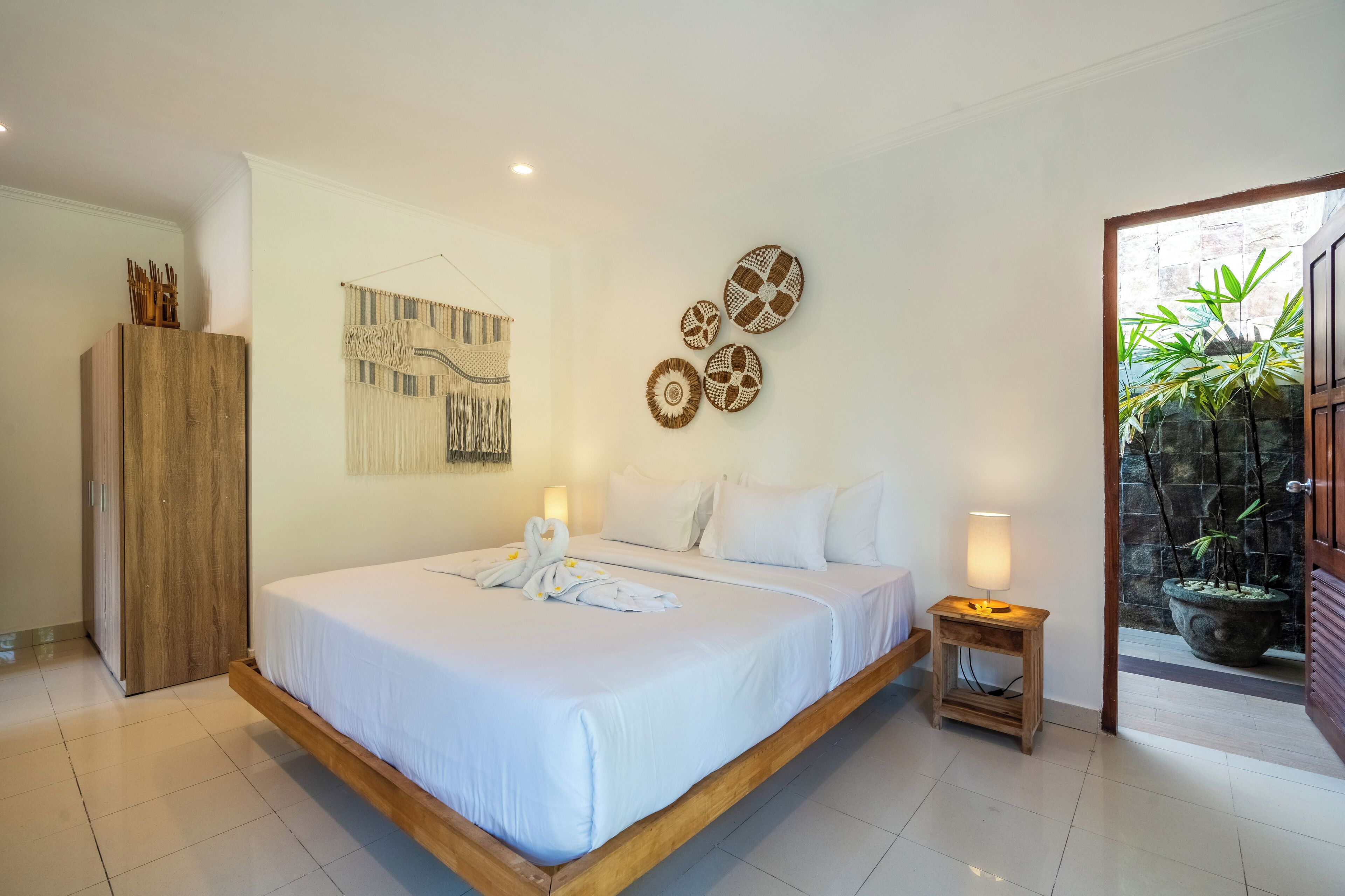 A bedroom with a large bed, a wooden wardrobe, a wall-mounted tapestry, and a potted plant near the entrance.