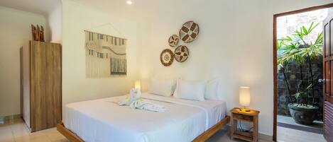 A bedroom with a large bed, a wooden wardrobe, a wall-mounted tapestry, and a potted plant near the entrance.