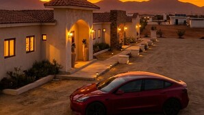 A red car parked in a desert-like setting with a sunset sky, a building with a tiled roof, and a clear view of the mountains in the background. - Baja Toscana Rancho Olivares (Valle de Guadalupe)