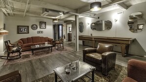 A spacious living area with a brown leather sofa, a coffee table, and a billiard table. There are multiple chairs and a small table with a glass top. The room has a high ceiling with exposed pipes and a hanging light fixture. - The Dillinger Room @ The Bank (East Aurora)