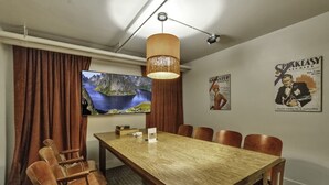Een vergaderruimte met een houten tafel, zes stoelen, een televisie waarop een berglandschap wordt getoond, een hanglamp en twee posters aan de muur.