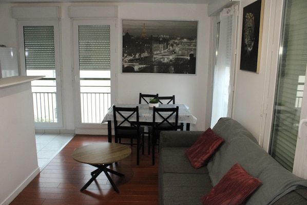 A living room with a gray sofa, a wooden coffee table, and a dining table with chairs. There is a framed picture on the wall and a window with blinds.