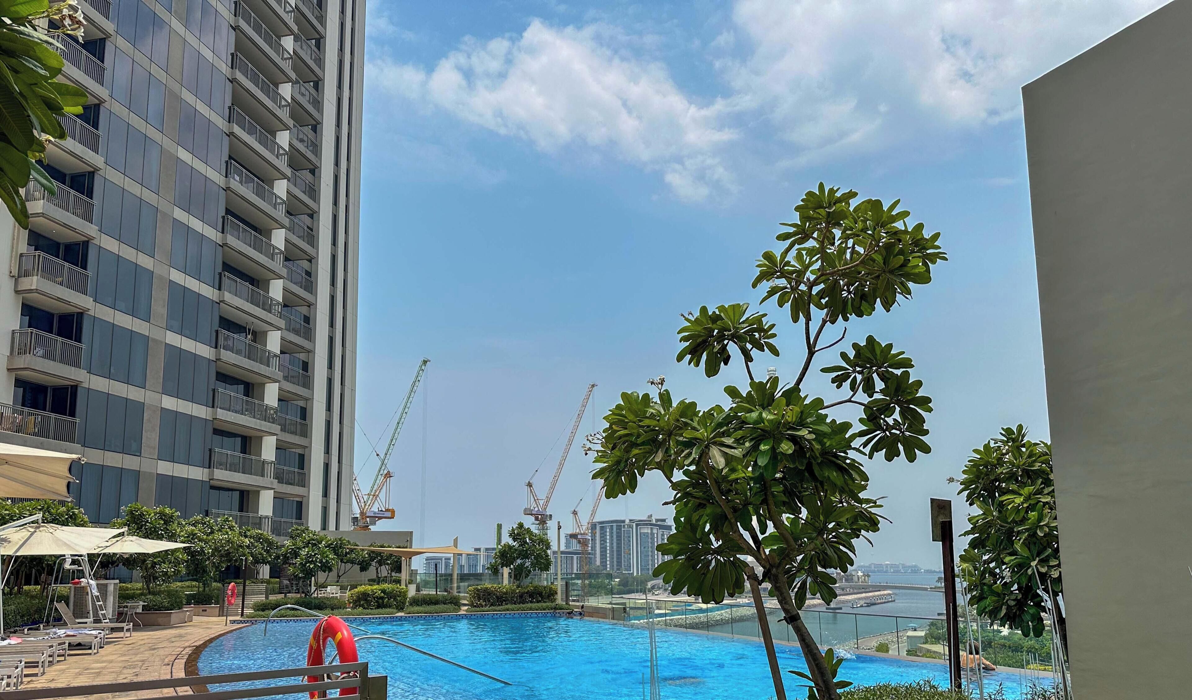 A swimming pool with a lifebuoy, surrounded by lounge chairs and umbrellas, with a view of a high-rise building and construction cranes in the background.