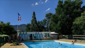 A swimming pool with clear blue water, surrounded by trees and a building with a flag.