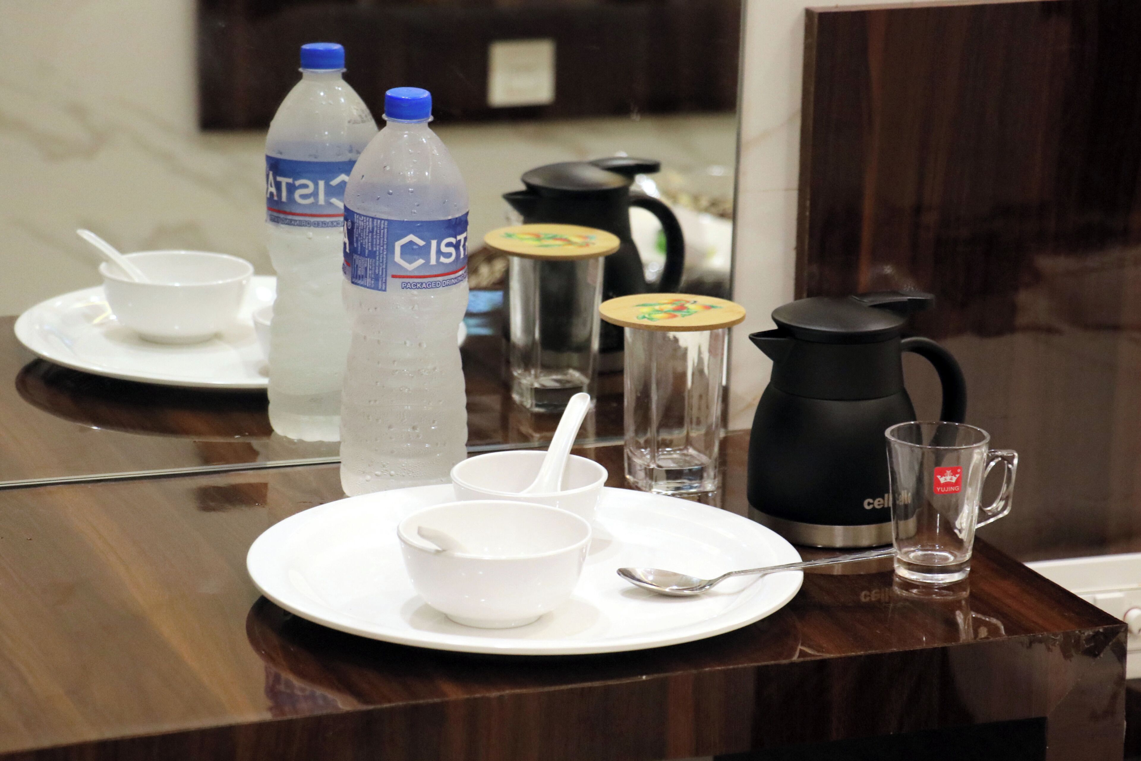 A hotel room table set with a water bottle, two bowls, a spoon, a coffee pot, and a glass.