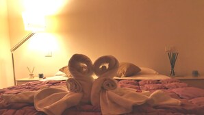 A bed with a heart-shaped towel arrangement, a bedside lamp, and a nightstand with a cup and reed diffuser.
