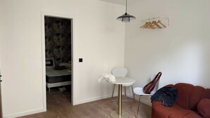 Un salon avec un canapé marron, une petite table ronde et une chaise blanche. On y trouve également un porte-manteau sur lequel sont accrochés des vêtements.