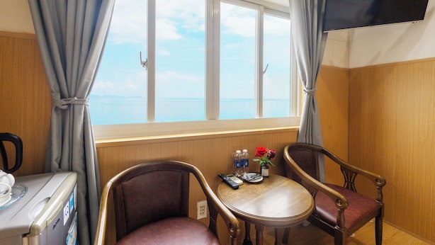Une chambre d’hôtel avec une grande fenêtre, deux chaises, une petite table avec une télécommande, une bouteille et un vase avec une fleur.