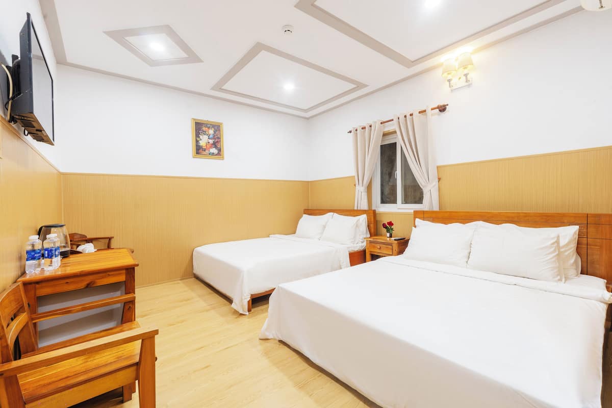 A hotel room with two single beds, wooden furniture, a flat-screen TV, a small table with a vase, and a window with curtains.