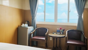 Une chambre d’hôtel comprenant un lit, deux chaises, une petite table avec une bouilloire et des tasses, un réfrigérateur et une fenêtre donnant sur la mer.