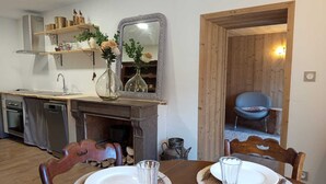 A dining area with a wooden table set for two, a kitchen with a built-in oven and dishwasher, and a mirror above the fireplace. - Au Mont Chablis (Champagnole)