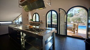 A modern kitchen with a central island, a built-in oven, and a large window overlooking a pool area. - Spacious 4BR Villa w/ AC, Private Pool, BBQ & Sea Views – Costa Brava (Sant Feliu de Guíxols)