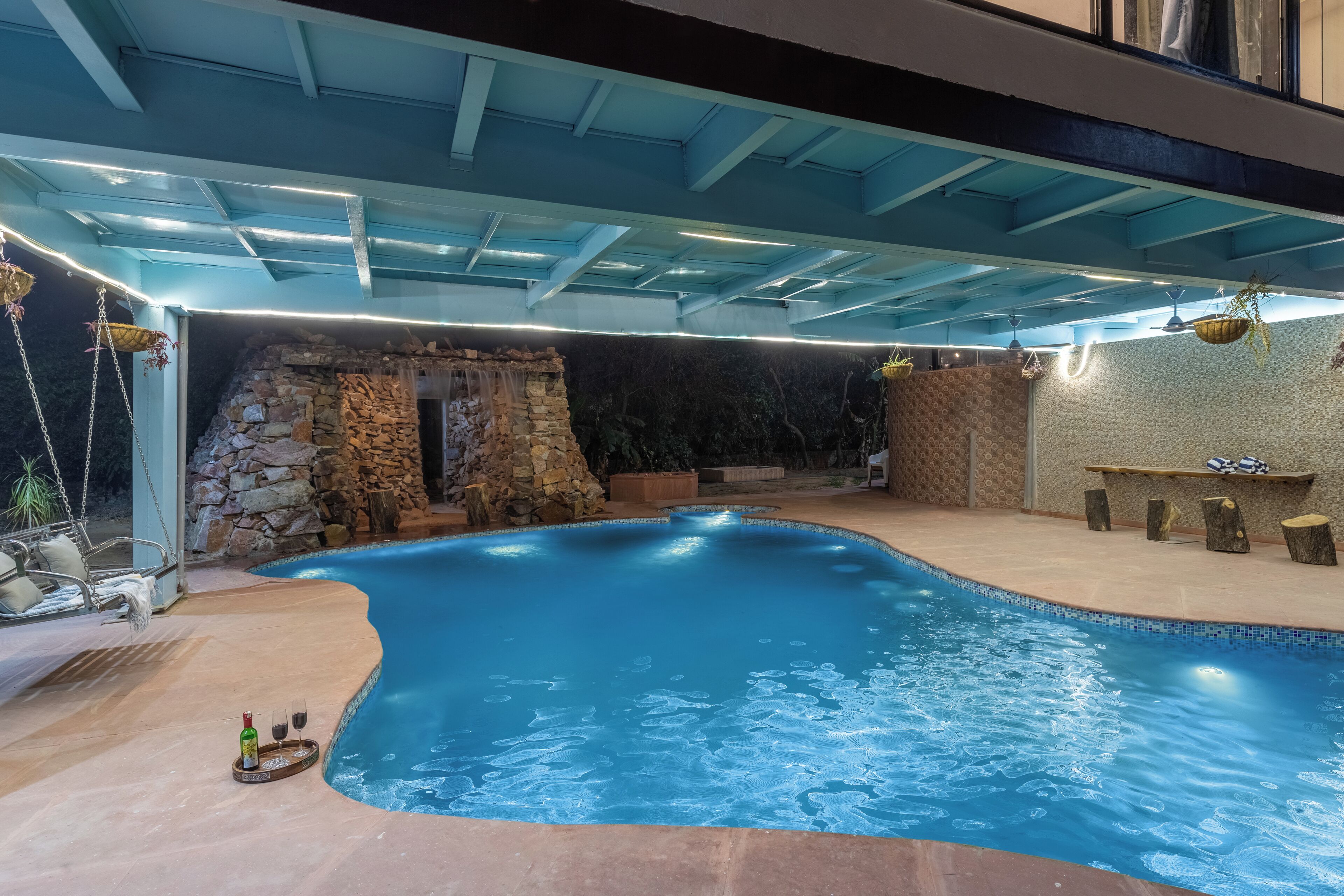 En swimmingpool med en stenvæg og hængeplanter, en træterrasse og et overdækket område med et blåt loft.