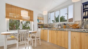 A kitchen with wooden cabinets, a dining table with chairs, a window with blinds, and a shelf with kitchenware. - Holiday Home 'Casa Azul' with Sea View, Private Terrace & Wi-Fi (Las Palmas)